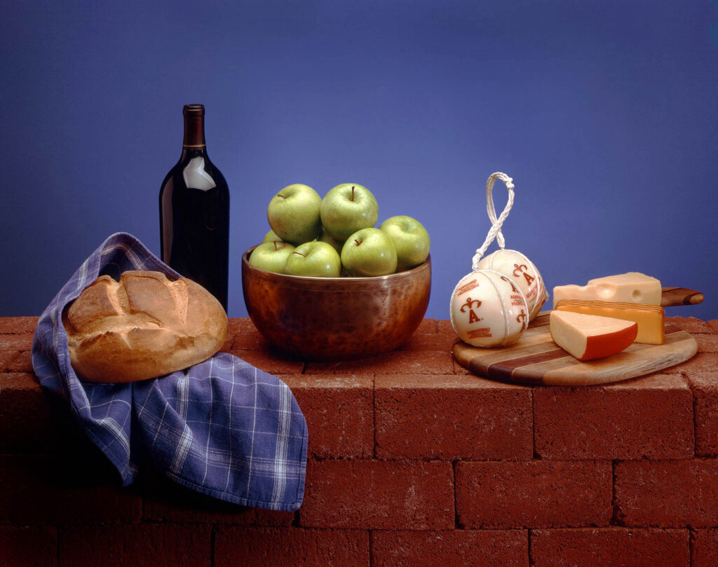A Still-life of pippin apples, cheeses and fresh baked bread with a bottle of red wine.  Still life photography at the Eagle Visions studio.