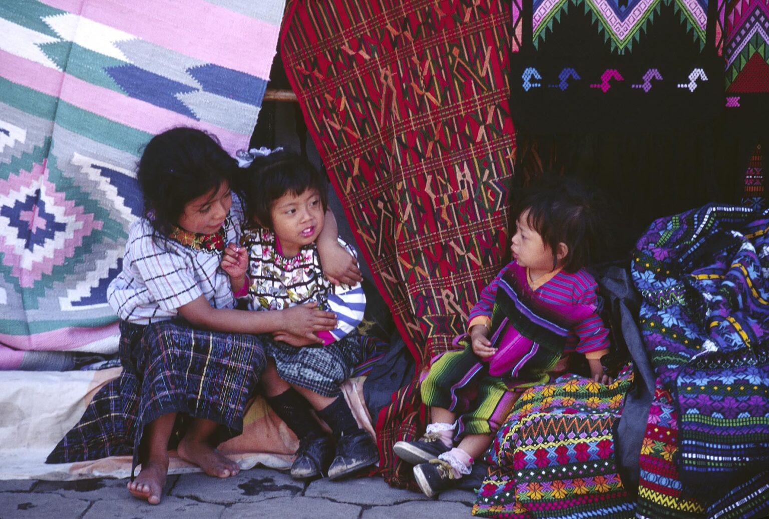 TZUTUJIL CHILDREN surrounded by handmade traditional TEXTILES for sale in the MARKETPLACE - SANTIAGO ATITLAN, GUATEMALA