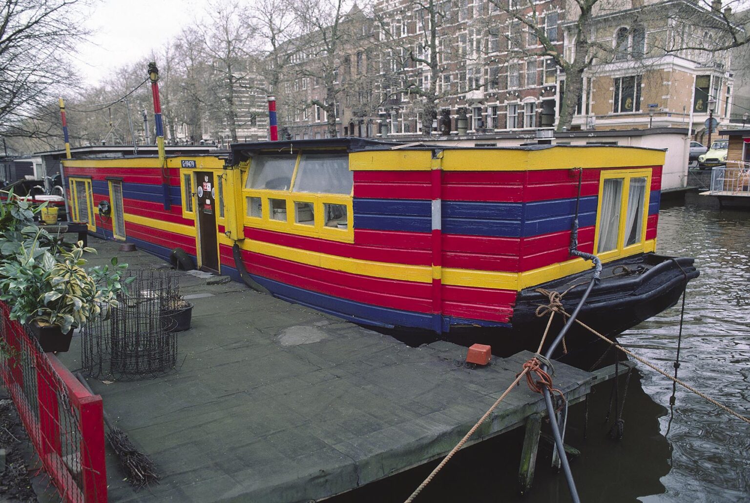 A HOUSEBOAT with a beautiful red, yellow & blue paint job is docked on a CANAL in AMSTERDAM