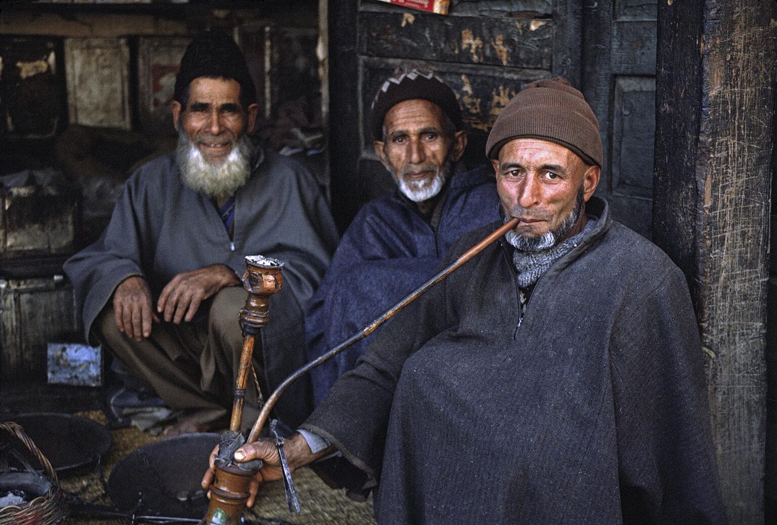 MUSLIM KASHMIRI MEN smoke a HUKKAH (hookah) in front of a small STORE - KASHMIR, INDIA