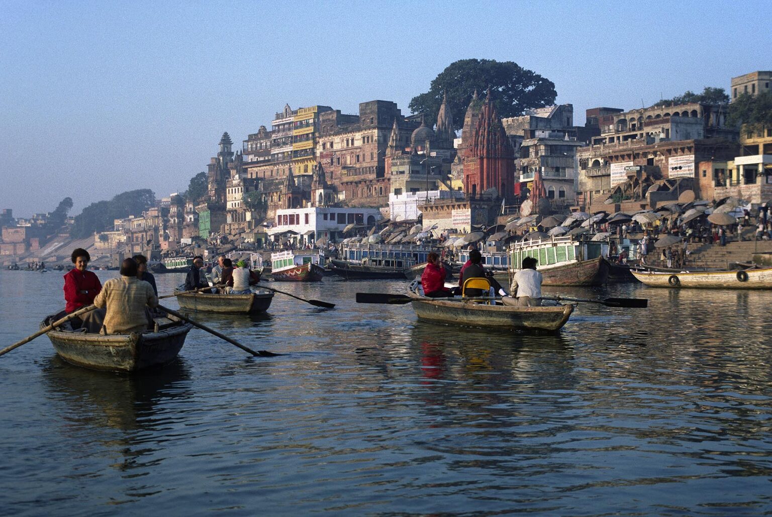 TOURISTS in ROWBOATS pass by a HINDU TEMPLE on the banks of the GANGES RIVER - VARANASI (BENARES), INDIA
