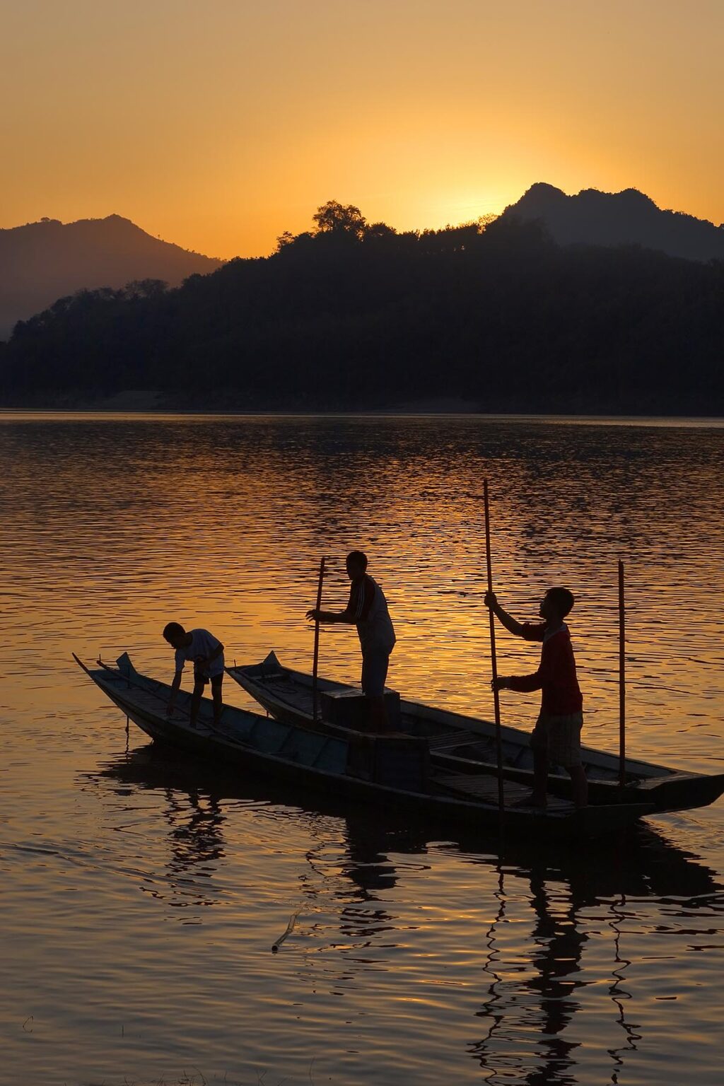 The sun sets over a hill on the Mekong River silhouetting Laotians polling their river boat as it runs through  - LUANG PROBANG,LAOS