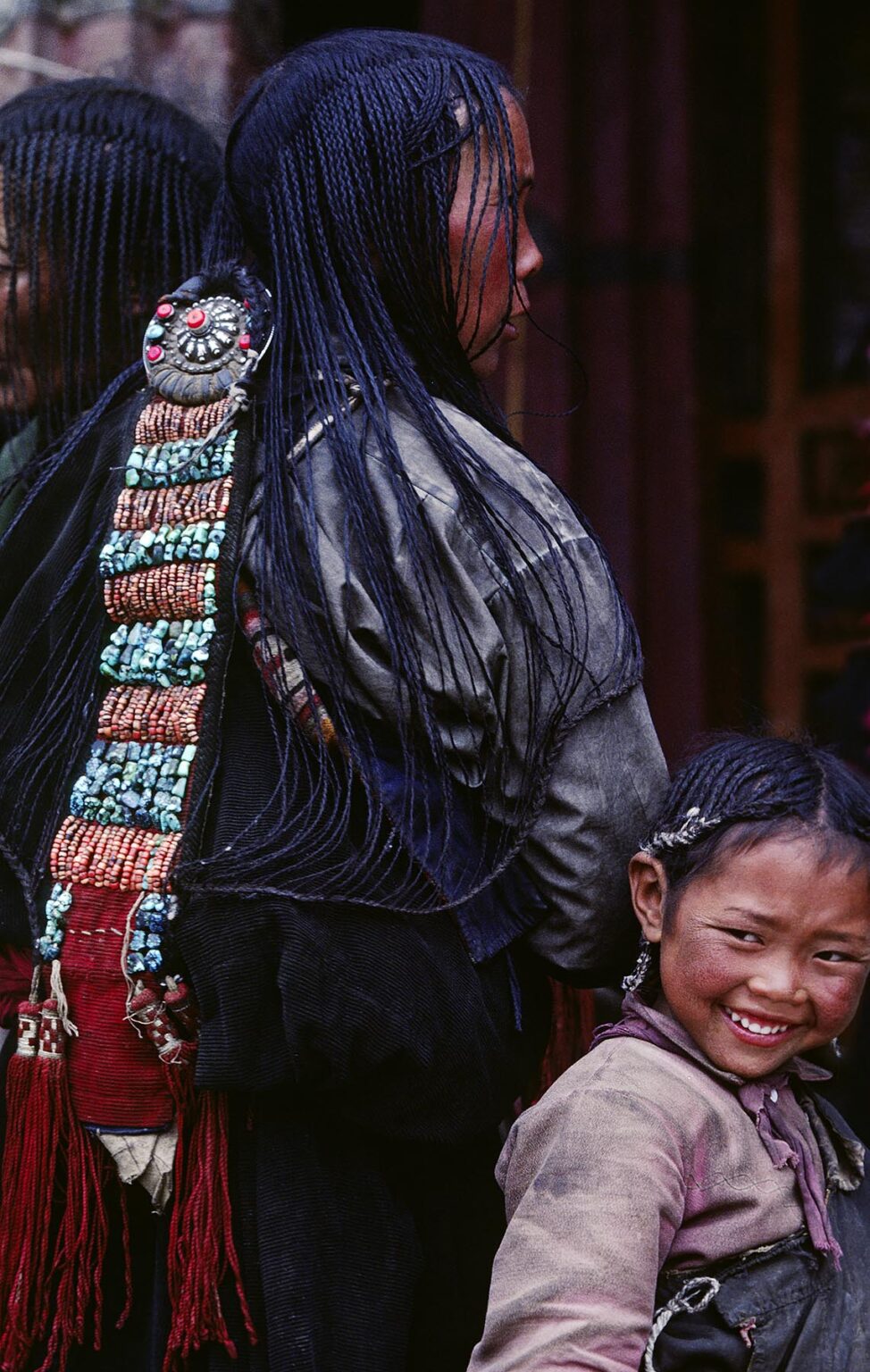 Young girl stands behind her mothers 108 braids & beautiful coral & turquoise hair piece - Sera Monastery, Lhasa, Tibet