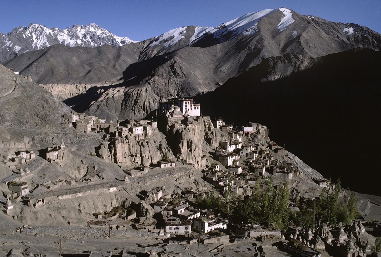 LAMAYURU'S chortens, fallow fields, village, and gompa (monastery), with HIMALAYAN PEAKS in background - LADAKH, INDIA