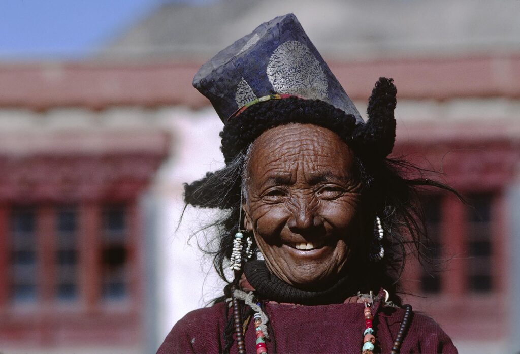 SMILING LADAKHI WOMAN wearing chuba, SEED PEARL earrings, coral and turquoise necklace, rosary and hat - LADAKH, INDIA