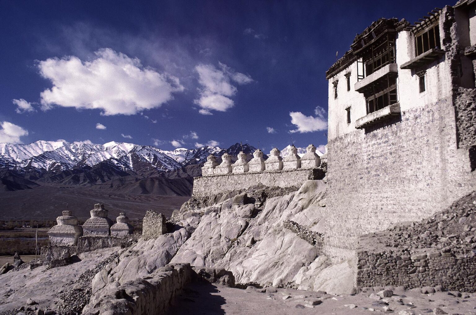 CHORTENS and the buildings of SHEY GOMPA (monastery), sit beneath HIMALAYAN PEAKS - LADAKH, INDIA