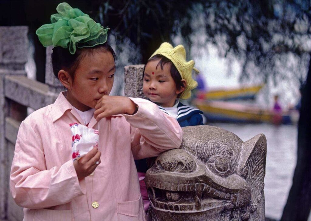 A CHINESE GIRL drinks from a packet while her SISTER sits on a STONE carved SNOW LION in KUNMING - YUNNAN, CHINA