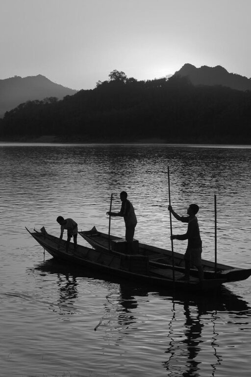 The sun sets over a hill on the Mekong River silhouetting Laotians polling their river boat as it runs through - LUANG PROBANG, LAOS